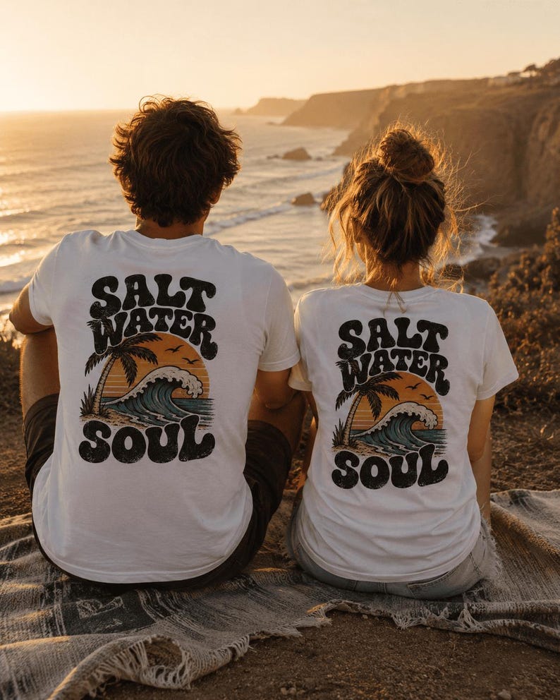 May include: Two white t-shirts with the text "SALT WATER SOUL" on the back, featuring a palm tree, waves, and a sunset design. The shirts are worn by two people sitting on a blanket, overlooking the ocean at sunset.
