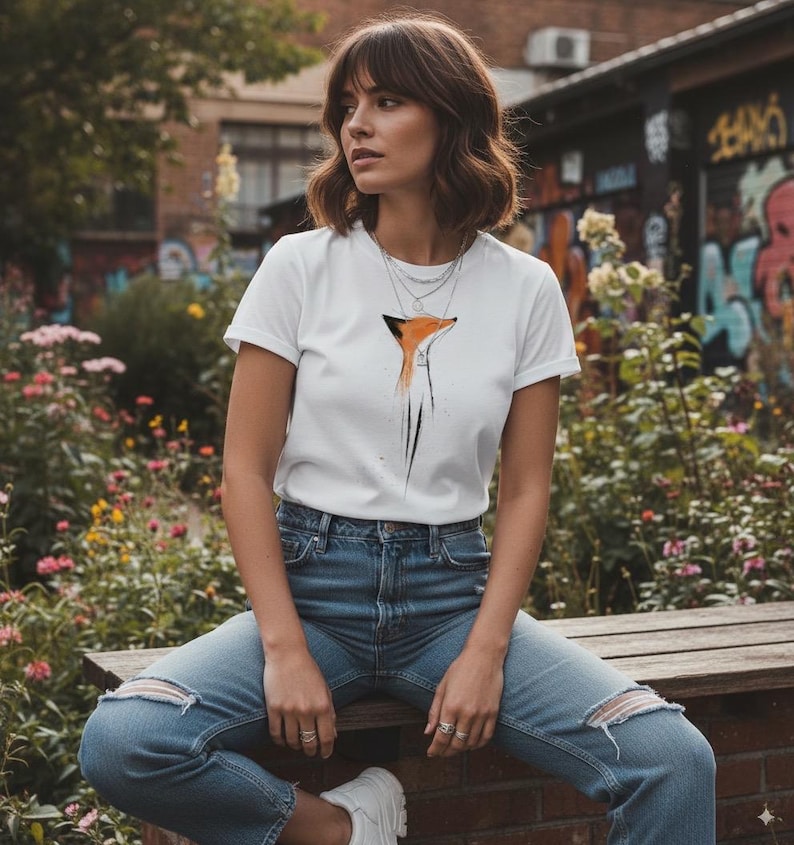 May include: A white short-sleeved t-shirt featuring a stylised orange and black fox graphic on the chest, paired with blue denim jeans that have distressed knee detailing. The t-shirt has a crew neckline and is worn with layered silver necklaces.