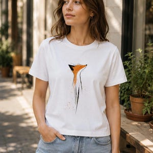 May include: A white short-sleeved t-shirt featuring a stylised graphic illustration of a fox head and neck in orange and black ink-wash style. The design is centred on the chest, showcasing a minimalist artistic depiction of the animal.