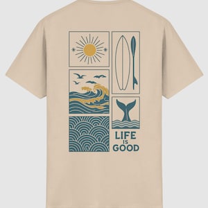 Pu&ograve; includere: Una t-shirt beige a maniche corte con una grafica a pi&ugrave; pannelli sul retro. Il design include un sole, onde oceaniche con uccelli, una tavola da surf, una coda di balena e un motivo a onde, con il testo "LIFE IS GOOD" stampato in blu scuro.
