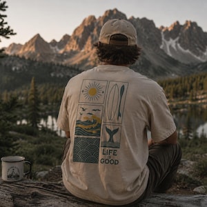 Pu&ograve; includere: Una t-shirt beige a maniche corte con una grafica sul retro composta da quattro pannelli rettangolari raffiguranti un sole, una tavola da surf, onde oceaniche e una coda di balena, con il testo "LIFE IS GOOD" stampato sotto la grafica.
