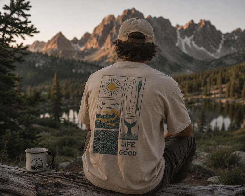 Pu&ograve; includere: Una t-shirt beige a maniche corte con una grafica sul retro composta da quattro pannelli rettangolari raffiguranti un sole, una tavola da surf, onde oceaniche e una coda di balena, con il testo "LIFE IS GOOD" stampato sotto la grafica.