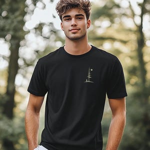 Op de afbeelding: Zwart T-shirt met ronde hals en een klein, minimalistisch ontwerp van een boom, maan en figuur op de linkerborst. Het shirt is gemaakt van zacht materiaal en wordt buiten gedragen.