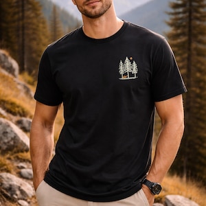 Könnte beinhalten: Schwarzes T-Shirt mit einer kleinen Grafik von Bäumen, einer Sonne und einem Fuchs. Das Shirt wird mit khakifarbenen Cargohosen kombiniert. Eine schwarze Uhr ist sichtbar. Der Hintergrund zeigt eine Berglandschaft.