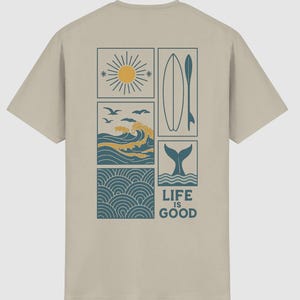 Pu&ograve; includere: Una t-shirt beige a maniche corte con una grafica sul retro composta da quattro riquadri rettangolari. I riquadri mostrano un sole giallo, una tavola da surf con pagaia, onde oceaniche con uccelli e una coda di balena. Il testo LIFE IS GOOD &egrave; stampato sotto.