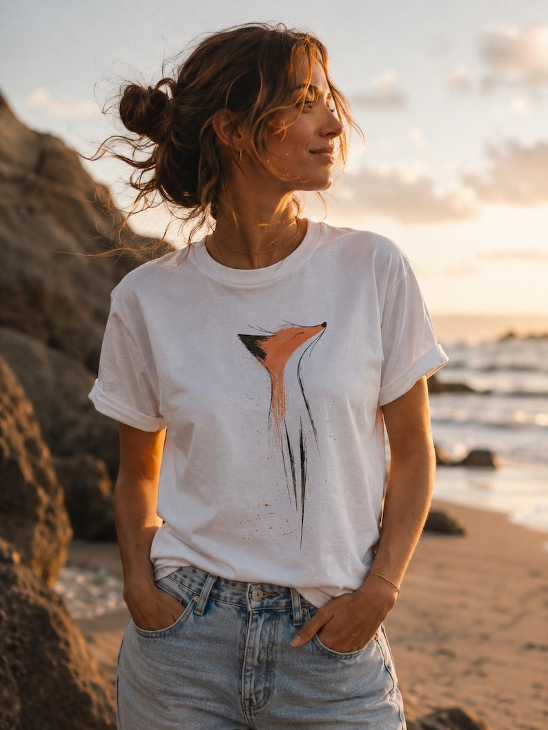 May include: A white short-sleeved t-shirt featuring a stylised graphic illustration of a fox head and neck in orange and black ink-wash style. The design is centred on the chest, showcasing a minimalist artistic depiction of the animal.