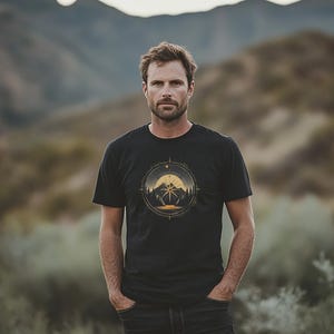 Könnte beinhalten: Schwarzes T-Shirt mit einem kreisförmigen Grafikdesign, das einen Berg und eine Sonne zeigt. Das Design verwendet Gelb- und Brauntöne. Die Person trägt eine schwarze Jeans und steht draußen.