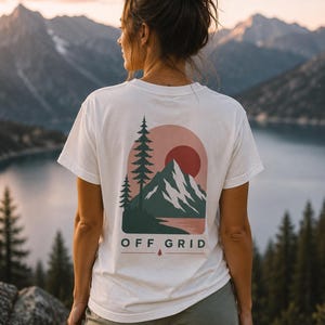 Puede incluir: Camiseta blanca con un gráfico de montaña y puesta de sol en la espalda. El diseño presenta una cordillera, árboles y un sol rojo, con las palabras "OFF GRID" debajo. La camiseta se usa al aire libre con un telón de fondo escénico de montaña y lago.