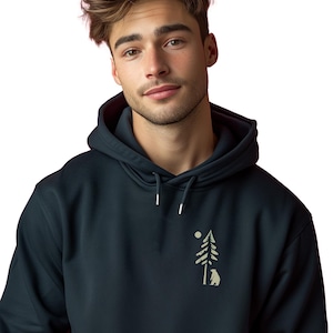 Op de afbeelding: Een marineblauwe hoodie met een trekkoord. De hoodie heeft een kleine afbeelding van een boom, zon en beer in een lichtbruine kleur op de borst. De persoon op de afbeelding draagt de hoodie.