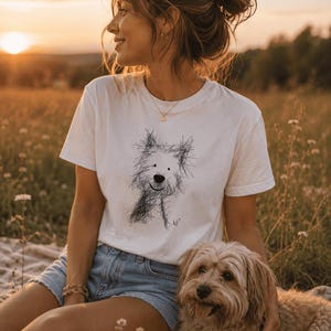 Könnte beinhalten: Ein weißes Kurzarm-T-Shirt mit einer schwarzen, skizzenhaften Zeichnung eines struppigen Hundes auf der Vorderseite. Die Grafik zeigt einen stilisierten Hund mit unordentlichen Linien, einer einfachen Nase und einem kleinen Lächeln auf der Brust.