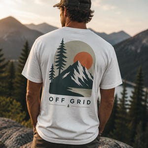 Op de afbeelding: Een wit t-shirt met een berg- en zonsondergangafbeelding op de rug. De afbeelding toont een bergketen, een ondergaande zon en dennenbomen. De tekst "OFF GRID" staat eronder gedrukt. De persoon draagt een groene pet.