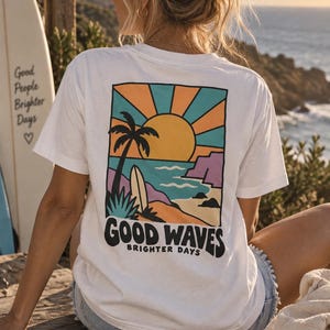 Könnte beinhalten: Ein weißes Kurzarm-T-Shirt mit einer rechteckigen Grafik auf der Rückseite. Die Illustration zeigt eine Strandszene mit einer Palme, einem Surfbrett und der Sonne. Der Text "GOOD WAVES" und "BRIGHTER DAYS" ist schwarz unter der Grafik gedruckt.