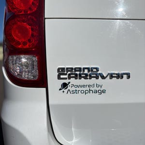 May include: Close-up of a white vehicle's rear with a red tail light and the chrome text "GRAND CARAVAN". Below, a black sticker reads "Powered by Astrophage" with a planet and star graphic.