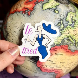 May include: A sticker with a cartoon illustration of a woman wearing a blue dress and a blue hat. The woman is holding a cigarette and the text "le tired" is written in pink below her. The sticker is placed on a globe.