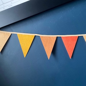 May include: A felt pennant banner with six triangular flags in shades of brown, yellow, orange, and red. The flags are strung together with a white string.