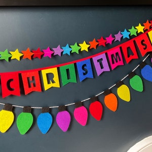 May include: A colorful felt Christmas banner with the word "CHRISTMAS" in white letters. The banner is strung with a red ribbon and is decorated with rainbow stars. Below the banner is a string of rainbow felt light bulbs.