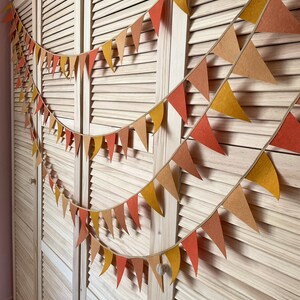 May include: A festive garland of felt triangle flags in shades of orange, yellow, and brown hangs on a wooden door. The garland is strung with twine and features a variety of sizes and shapes of flags.