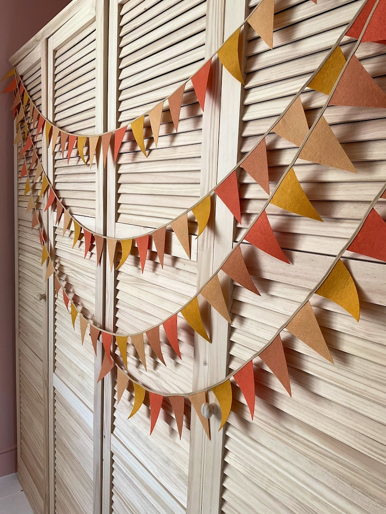 May include: A festive garland of felt triangle flags in shades of orange, yellow, and brown hangs on a wooden door. The garland is strung with twine and features a variety of sizes and shapes of flags.