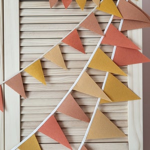 May include: A festive garland with triangular flags in shades of orange, yellow, and brown. The flags are strung together with white string and hang against a light wood background.