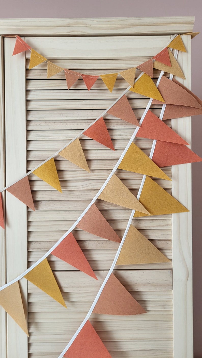 May include: A festive garland with triangular flags in shades of orange, yellow, and brown. The flags are strung together with white string and hang against a light wood background.