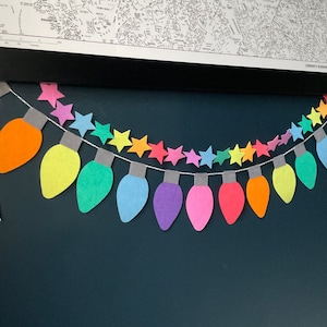 May include: A rainbow-colored paper garland of light bulbs and stars. The garland is strung on a white string and is hanging on a dark blue wall.