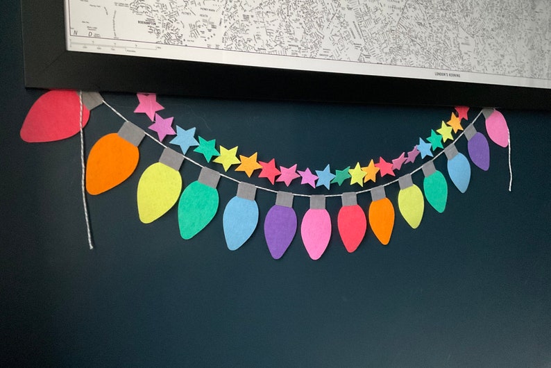 May include: A rainbow-colored paper garland of light bulbs and stars. The garland is strung on a white string and is hanging on a dark blue wall.