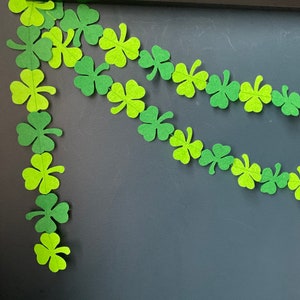 May include: A green shamrock garland with paper shamrocks strung together. The garland is hanging on a dark grey surface.