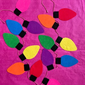 May include: A string of felt light bulbs in various colors, including pink, red, orange, yellow, green, blue, and purple, with black felt accents. The bulbs are strung together with white twine.