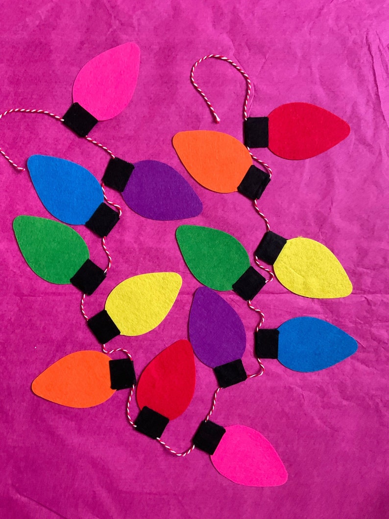 May include: A string of felt light bulbs in various colors, including pink, red, orange, yellow, green, blue, and purple, with black felt accents. The bulbs are strung together with white twine.