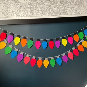 May include: A rainbow-colored felt garland of light bulbs, strung on a white string, hangs on a dark blue wall.