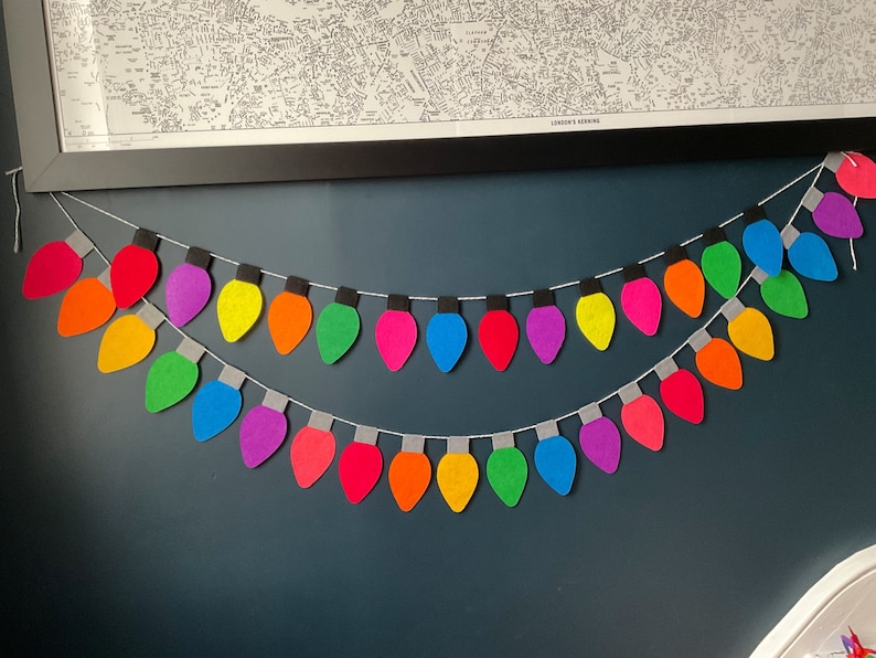 May include: A rainbow-colored felt garland of light bulbs, strung on a white string, hangs on a dark blue wall.