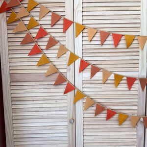 May include: A string of felt triangle flags in shades of orange, brown, and yellow hangs against a light wood background. The flags are strung together with twine.