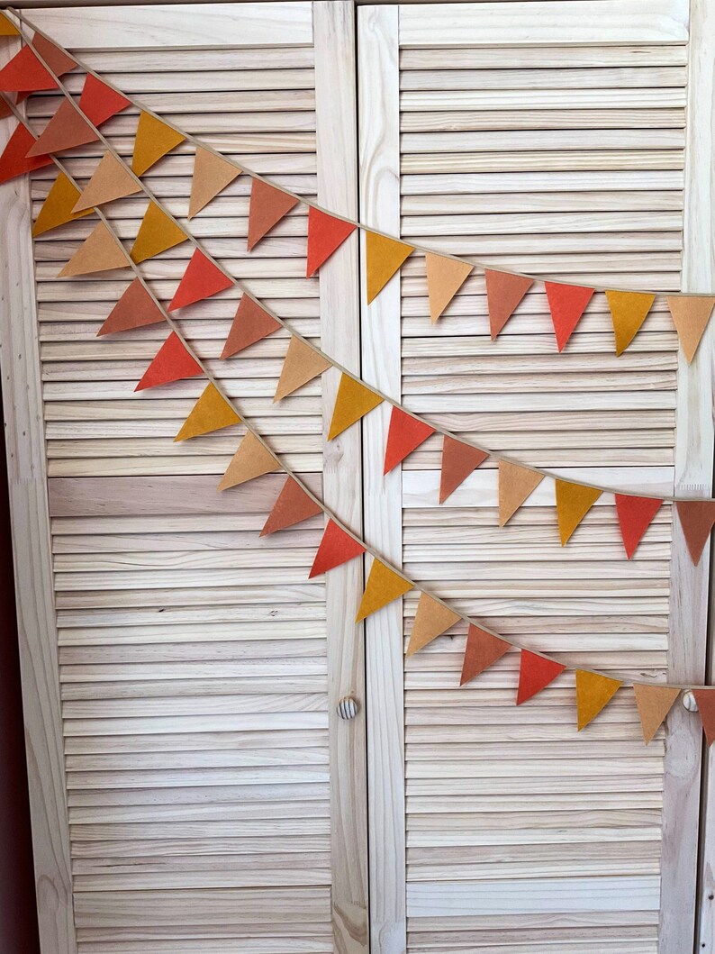 May include: A string of felt triangle flags in shades of orange, brown, and yellow hangs against a light wood background. The flags are strung together with twine.