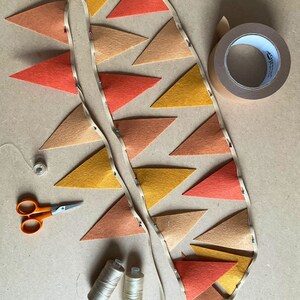 May include: A string of felt triangle flags in orange, yellow, and brown colors is being assembled. The flags are pinned together with safety pins and are being sewn onto a white ribbon. There are two spools of thread, a pair of scissors, and a roll of tape on the table.