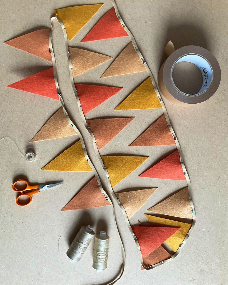 May include: A string of felt triangle flags in orange, yellow, and brown colors is being assembled. The flags are pinned together with safety pins and are being sewn onto a white ribbon. There are two spools of thread, a pair of scissors, and a roll of tape on the table.
