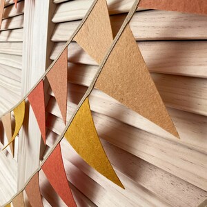 May include: A string of felt triangle flags in shades of orange, yellow, and brown, strung on a beige ribbon. The flags are hanging against a light wood background.