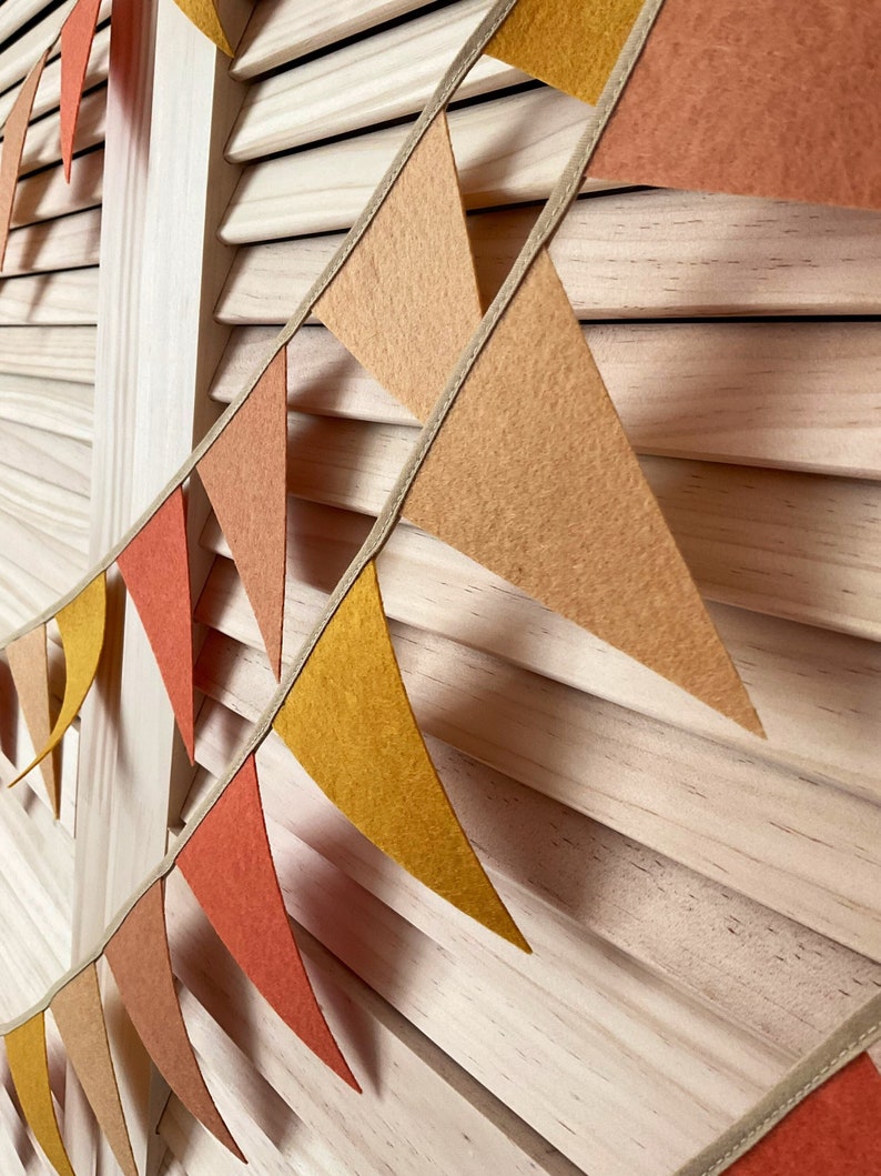 May include: A string of felt triangle flags in shades of orange, yellow, and brown, strung on a beige ribbon. The flags are hanging against a light wood background.