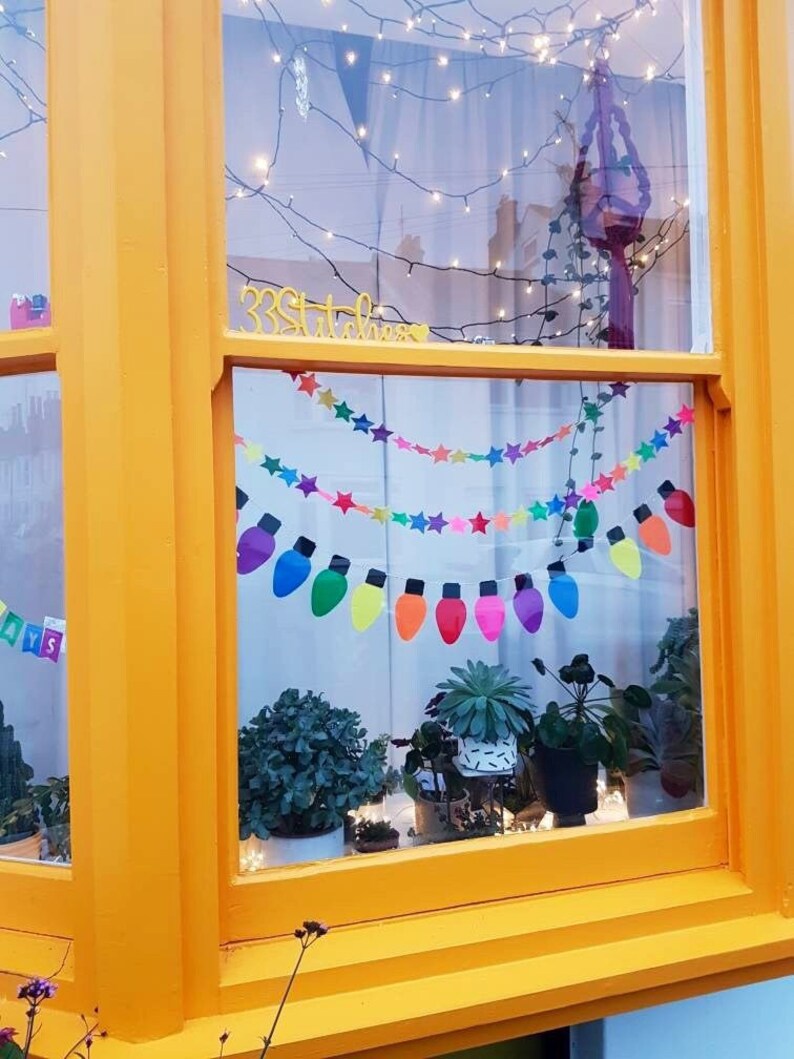 May include: A window with a yellow frame and a colorful string of lights with stars and light bulbs. The window is decorated with a sign that says "33 Stitches".
