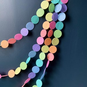 Peut inclure: Une guirlande arc-en-ciel faite de cercles en feutre. La guirlande est enfilée avec un fil blanc. Les cercles sont dans différentes nuances de rose, orange, jaune, vert, bleu et violet.