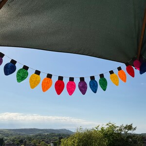 May include: A rainbow-colored string of felt light bulbs hangs from a patio umbrella against a blue sky. The light bulbs are in a variety of colors, including red, orange, yellow, green, blue, indigo, and violet.