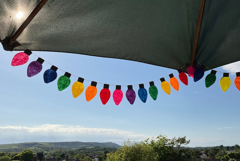 May include: A rainbow-colored string of felt light bulbs hangs from a patio umbrella against a blue sky. The light bulbs are in a variety of colors, including red, orange, yellow, green, blue, indigo, and violet.