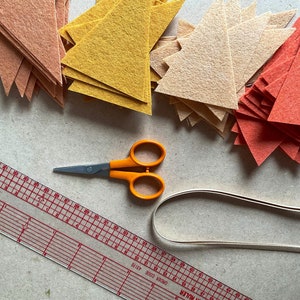 May include: A flat lay of craft supplies for making a felt banner. The image shows a ruler, scissors, a roll of white ribbon, and stacks of felt triangles in orange, yellow, and peach.