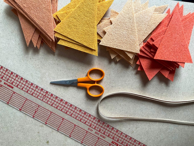 May include: A flat lay of craft supplies for making a felt banner. The image shows a ruler, scissors, a roll of white ribbon, and stacks of felt triangles in orange, yellow, and peach.