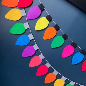 May include: A colorful felt garland with light bulb shapes in a rainbow pattern. The garland is strung on a white string and has gray felt circles between each light bulb.