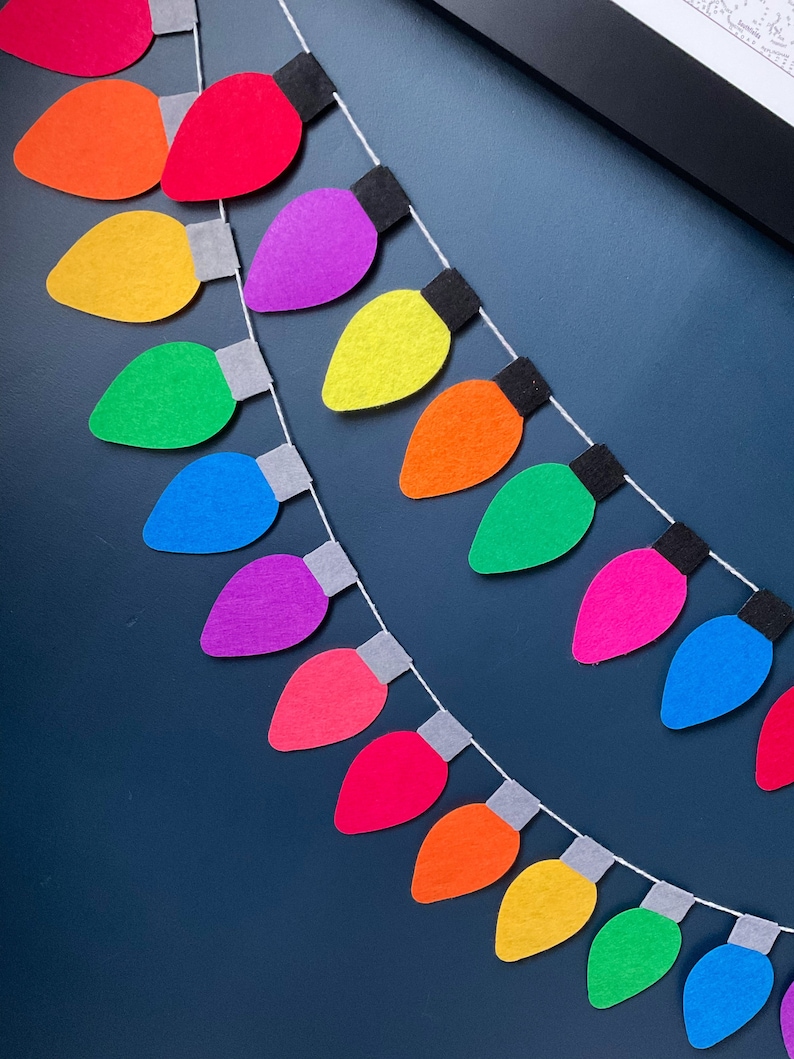 May include: A colorful felt garland with light bulb shapes in a rainbow pattern. The garland is strung on a white string and has gray felt circles between each light bulb.