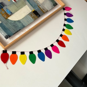 May include: A rainbow-colored felt garland of light bulbs, strung together with red thread. Each light bulb is a different color of the rainbow, starting with red and ending with purple.