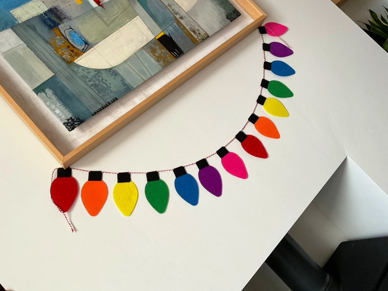 May include: A rainbow-colored felt garland of light bulbs, strung together with red thread. Each light bulb is a different color of the rainbow, starting with red and ending with purple.
