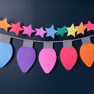 May include: A colorful felt garland with light bulbs and stars. The garland is made of felt and is strung together with white thread. The light bulbs are in various colors, including green, blue, purple, pink, red, orange, and yellow. The stars are in various colors, including yellow, orange, red, pink, purple, blue, and green.