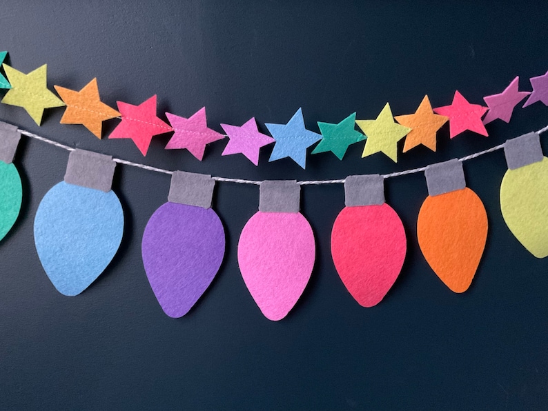 May include: A colorful felt garland with light bulbs and stars. The garland is made of felt and is strung together with white thread. The light bulbs are in various colors, including green, blue, purple, pink, red, orange, and yellow. The stars are in various colors, including yellow, orange, red, pink, purple, blue, and green.