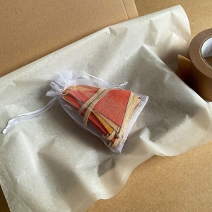 May include: A white tissue paper bag with a drawstring closure, filled with a stack of colorful paper pennants. The bag is sitting on a brown cardboard surface.